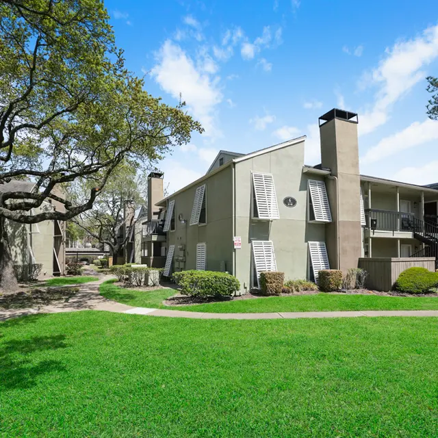 A view of a residential apartment complex surrounded by green lawns and trees under a clear blue sky.