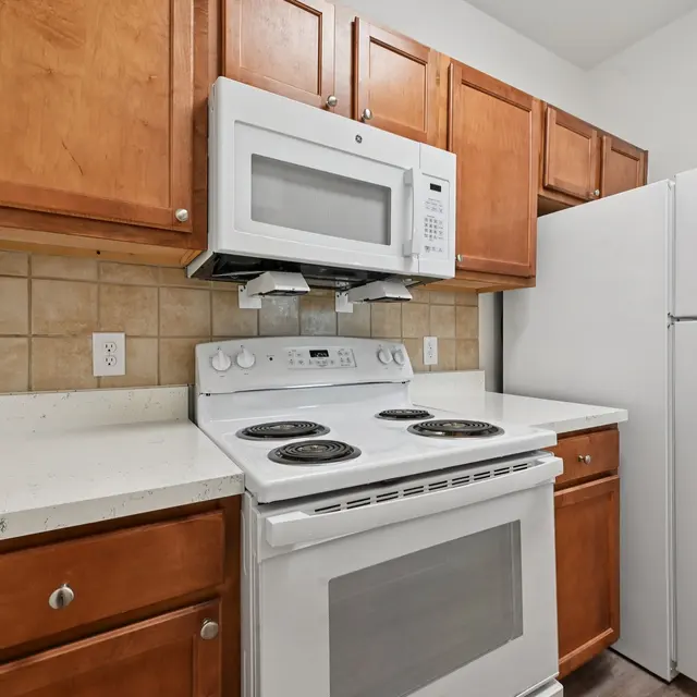 A modern kitchen featuring wooden cabinets, a white microwave and range hood, a white stove, and a refrigerator. The countertops are light-colored, and the backsplash is tiled.