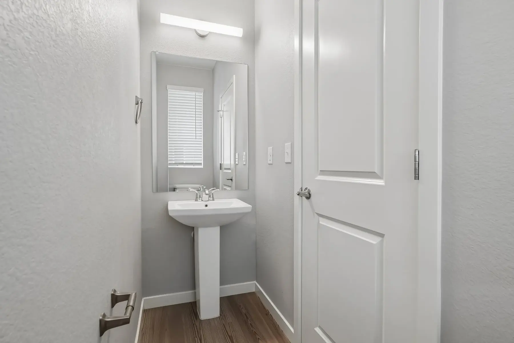 Compact Bathroom Overview A small bathroom with a pedestal sink and mirror, featuring light gray walls and wooden flooring.