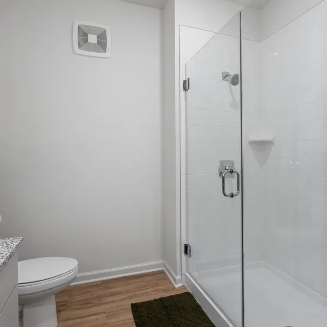 Modern Bathroom Interior A modern bathroom featuring a glass shower enclosure, a white toilet, and a vanity with a gray granite countertop. The walls are painted white, and there is a towel hanging by the vanity. A green bath mat lies on the floor, and a framed floral picture is hung on the wall.