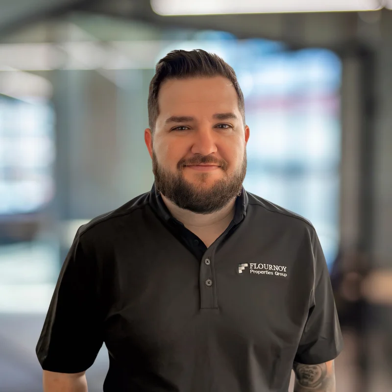 A man with a beard wearing a black polo shirt with a logo, standing indoors with a blurred background.