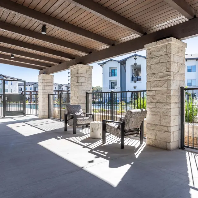 A covered patio area with two chairs and a small table, featuring stone columns and a view of modern apartment buildings in the background.