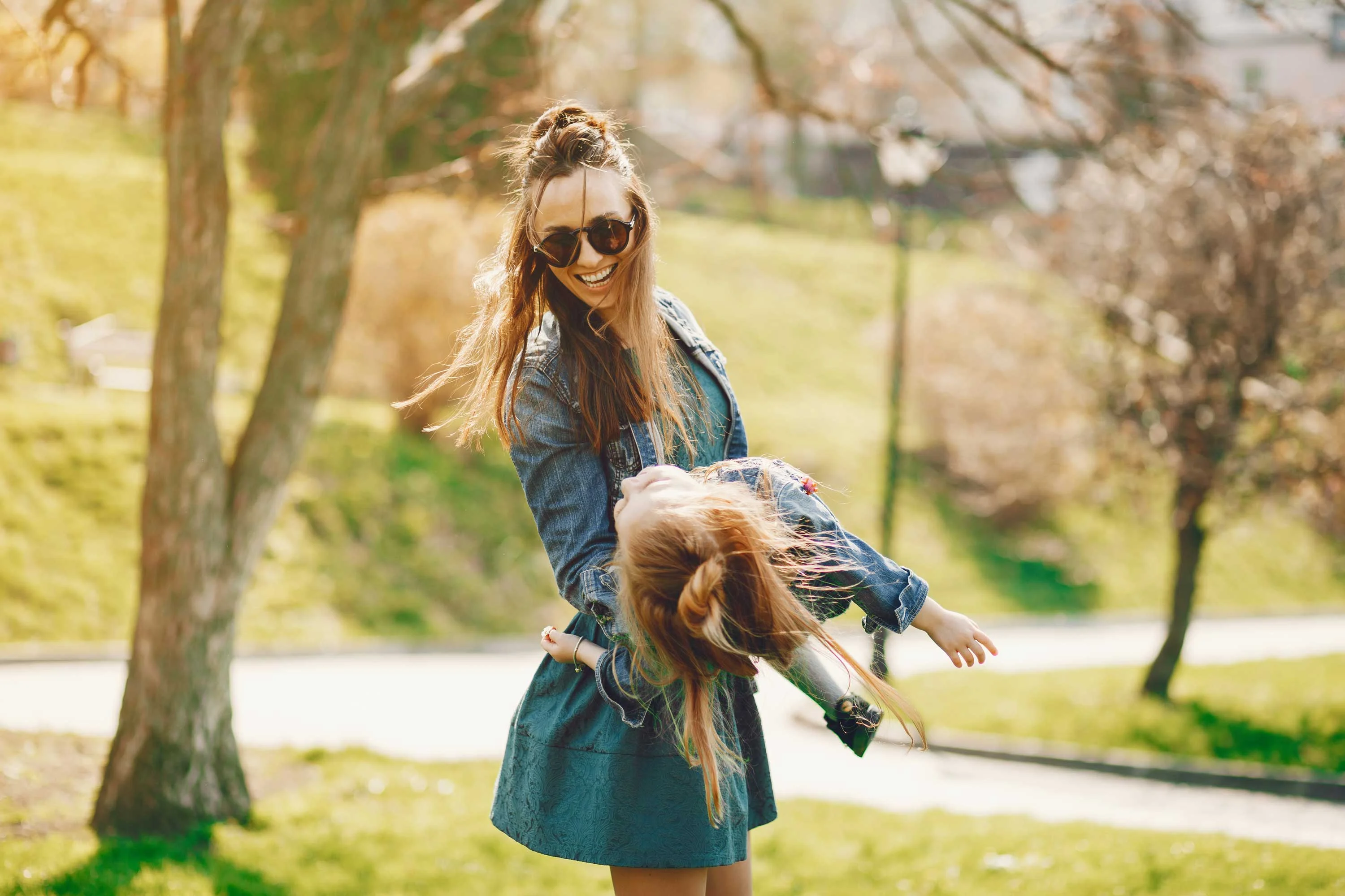 A young woman with sunglasses smiles while playfully holding a doll by its hair in a sunny park setting. The background features green grass and blurred trees, suggesting a joyful outdoor atmosphere.