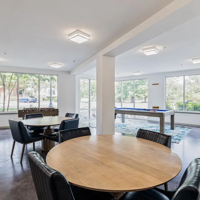 Modern Common Area A spacious and modern common area featuring a round wooden table surrounded by black chairs, with large windows allowing natural light. A ping pong table is visible in the background, and a blue armchair is placed near the windows.