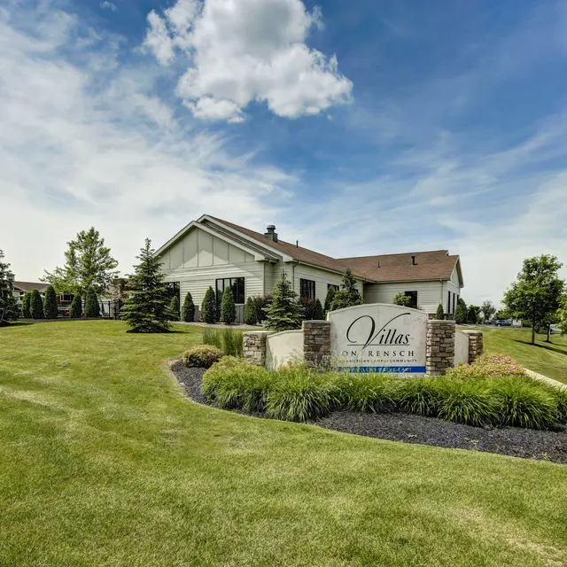 Exterior view of a residential building with a sign that reads 'Villas in Rich'. The landscape features green grass, trees, and shrubs under a blue sky with clouds.