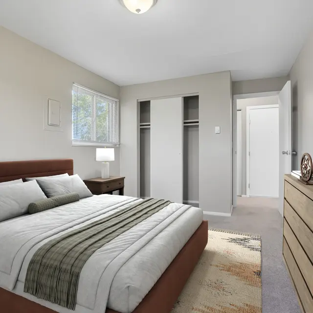 A modern bedroom featuring a queen-sized bed with gray bedding, wooden nightstands with lamps, a dresser, and a large closet. There is a window with natural light and a neutral color scheme throughout the room.