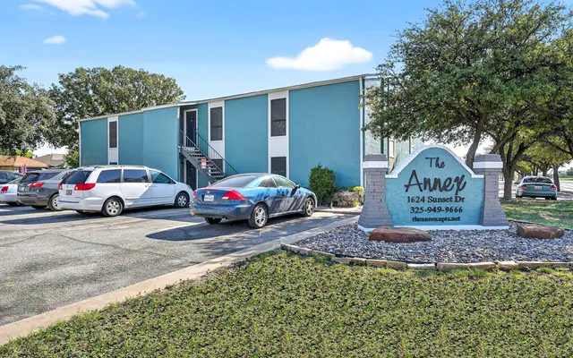 Exterior view of an apartment building named The Amer, featuring a parking area with several cars and landscaped grass.