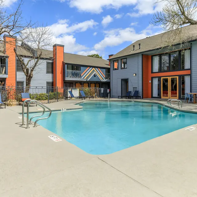 A clear swimming pool surrounded by lounge chairs and apartment buildings on a bright day with blue skies and scattered clouds.