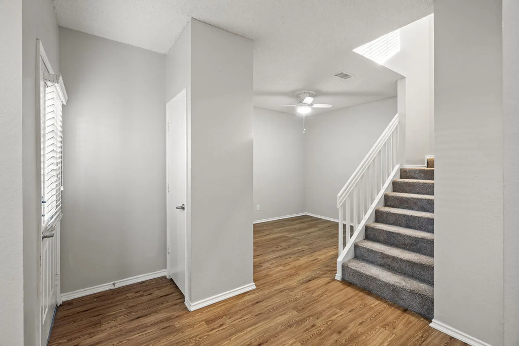 Interior Hallway with Staircase A spacious interior hallway leading to a staircase, with light grey walls and a wood-like floor. There is a ceiling fan in the corner and an open room visible to the right.