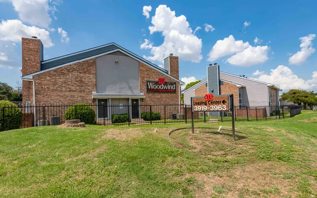Exterior view of the Woodwind Irving, showcasing two buildings with a sign for Woodwind and a learning center. The landscape is green with blue skies and scattered clouds.