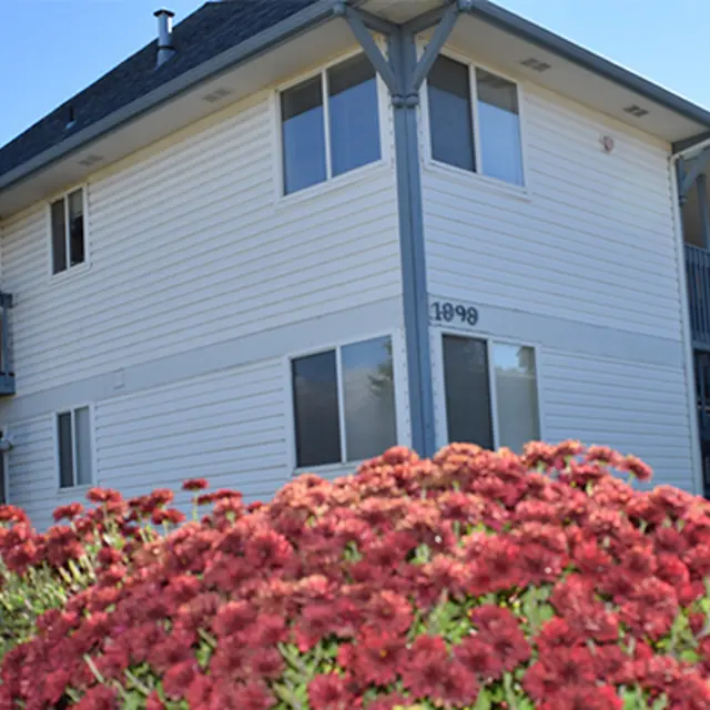 A two-story apartment building with white siding, featuring large windows and balconies, surrounded by red flowers in the foreground.