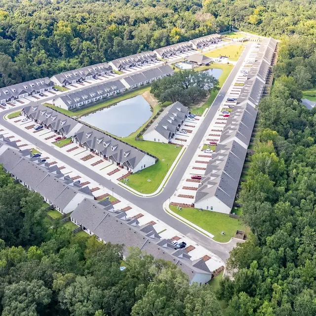 Aerial View of Residential Complex Aerial view of a residential complex surrounded by trees, featuring rows of houses and a small pond.