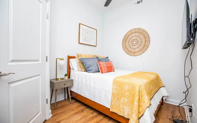 A small and cozy bedroom featuring a wooden bed with a yellow blanket, decorative pillows, a bedside table, and a wall-mounted TV.