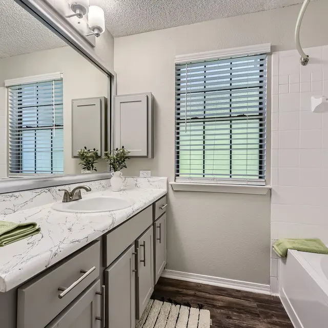 A modern bathroom featuring a marble countertop sink, gray cabinetry, and a bathtub. Natural light streams through a window with horizontal blinds.