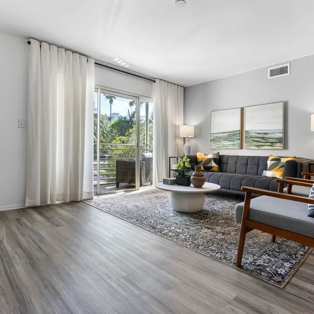 A modern living room featuring a comfortable gray sofa with yellow accents, a round coffee table, and stylish armchairs. Large windows with sheer curtains allow natural light, and there are decorative plants and artworks on the wall.
