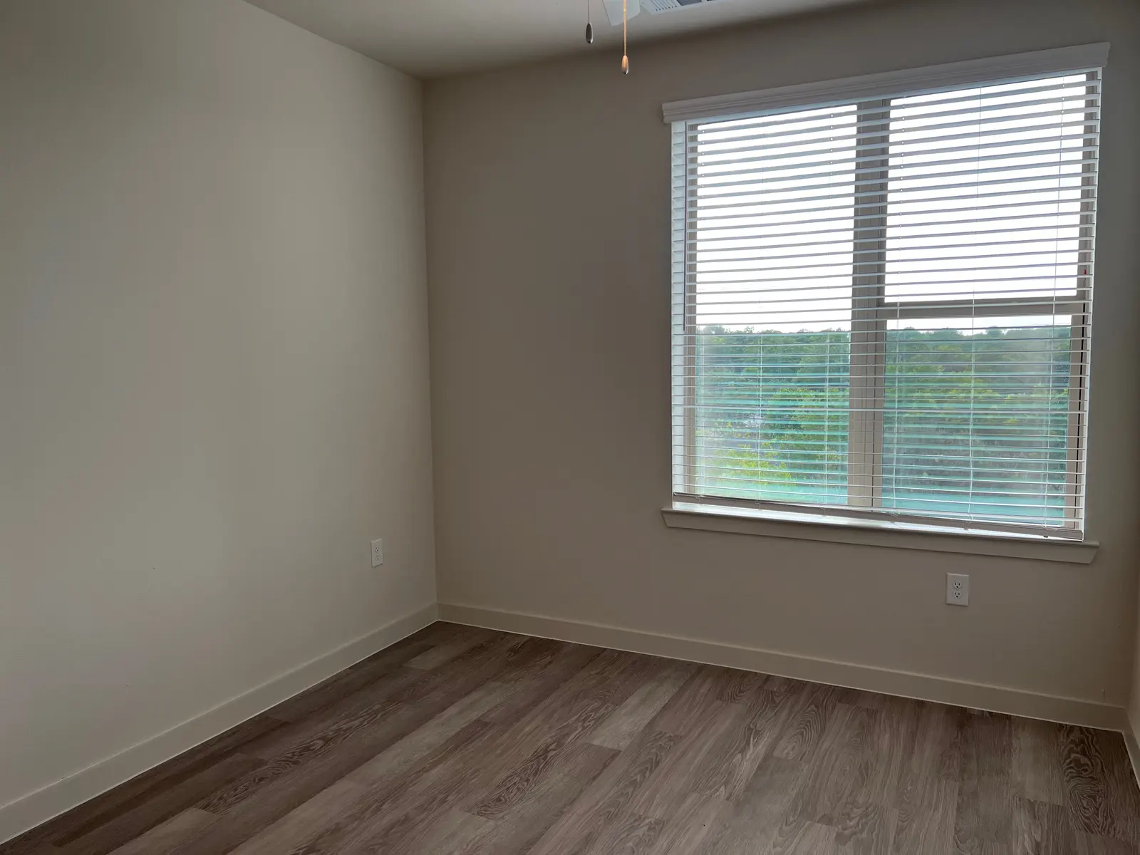 An empty room featuring a window with blinds, overlooking a green outdoor area.