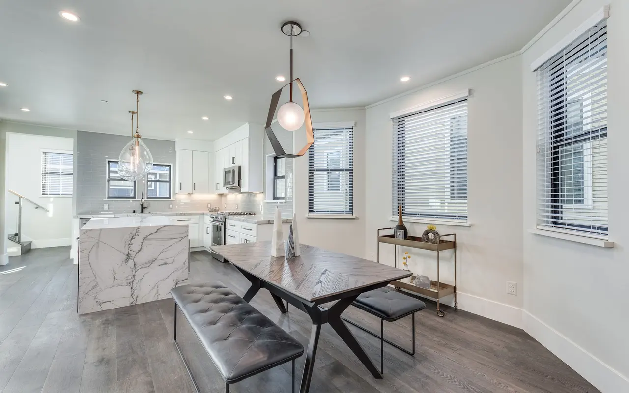 A modern kitchen and dining area featuring a marble tabletop dining table, black benches, pendant lighting, and large windows with blinds.