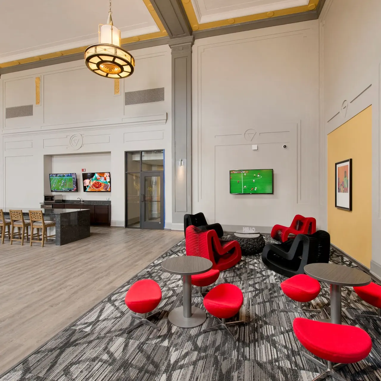 A spacious modern lounge area featuring red and black seating, a dining table, and screens displaying sports events on the walls.