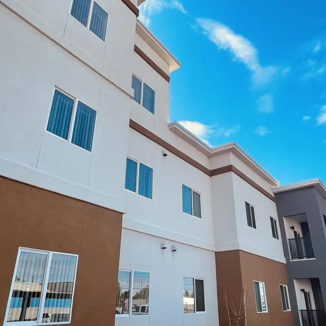 A modern multi-story apartment building featuring a mix of white and brown facades, large windows, and a clear blue sky in the background.