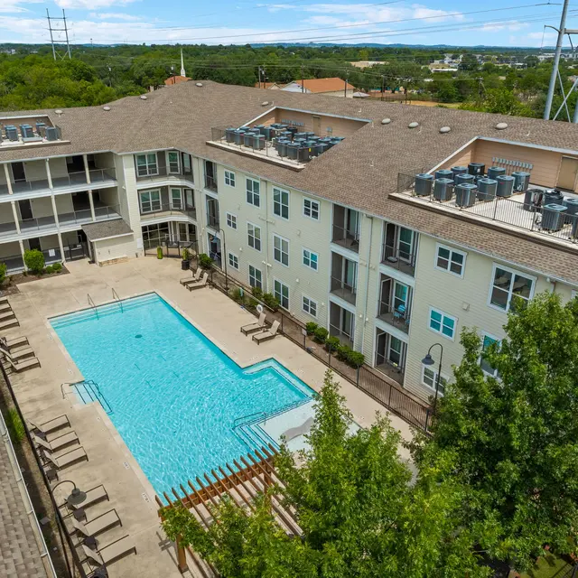 An aerial view of an apartment complex with a swimming pool in the center. The pool is surrounded by lounge chairs, and there are balconies with people relaxing on them. Lush green trees are visible around the area, and the sky is clear blue with a few clouds.