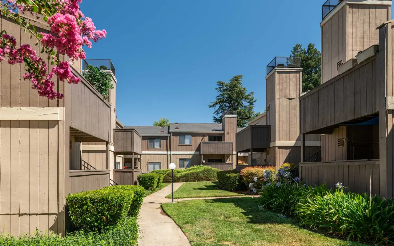 A well-maintained apartment complex with multiple brown buildings, green lawns, and blooming flowers under a clear blue sky.