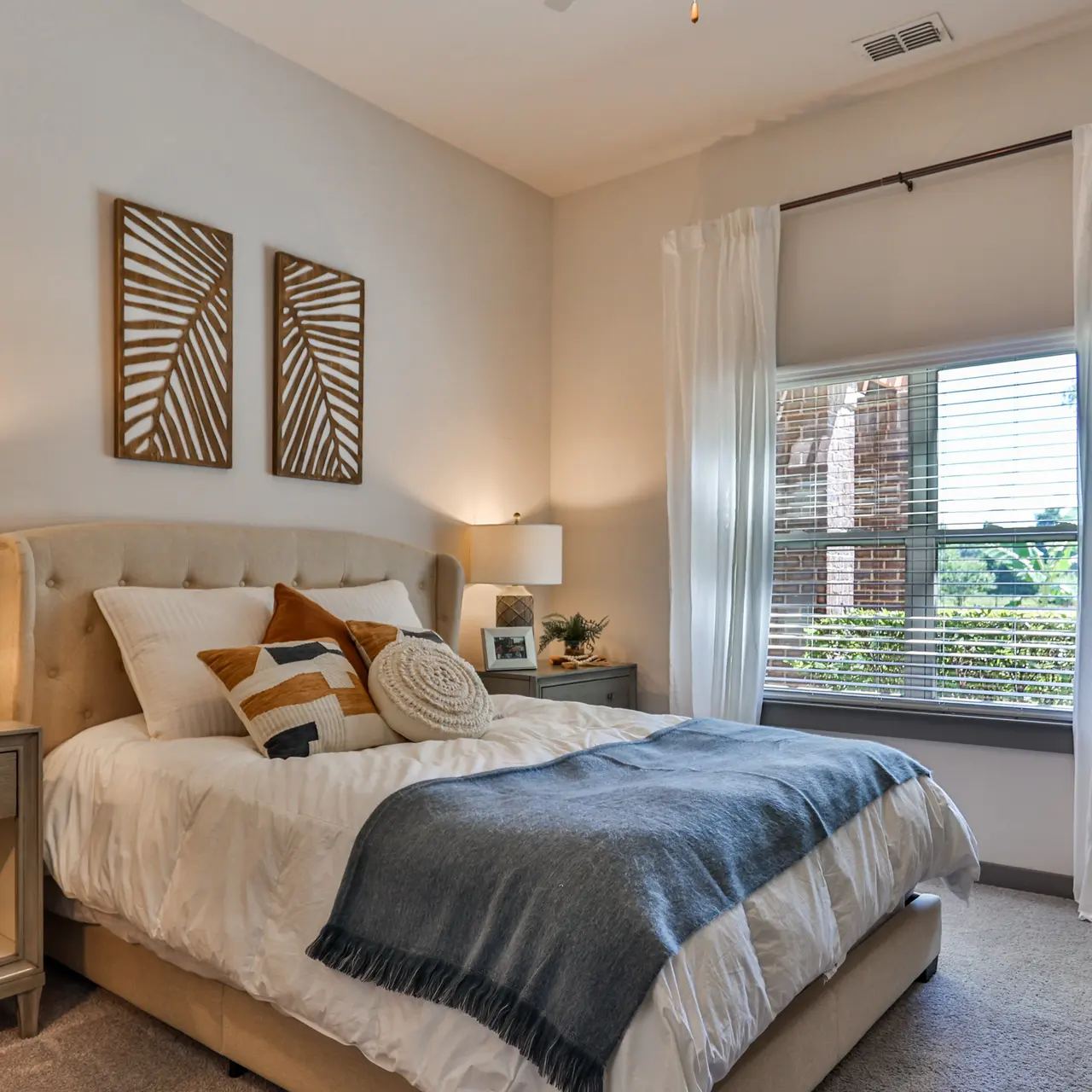 A cozy modern bedroom featuring a plush bed with decorative cushions, two wall art pieces, and natural light streaming in through a window adorned with sheer curtains.
