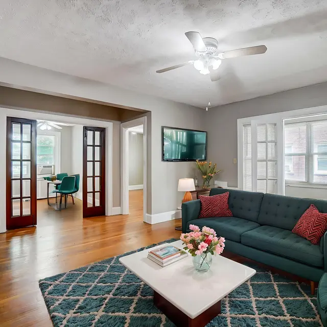 A stylish and modern living room featuring a dark blue sofa with decorative pillows, a white coffee table with a floral centerpiece, and a ceiling fan. In the background, glass French doors lead to a bright room with a green chair and white walls, while a mounted TV is visible on the wall.