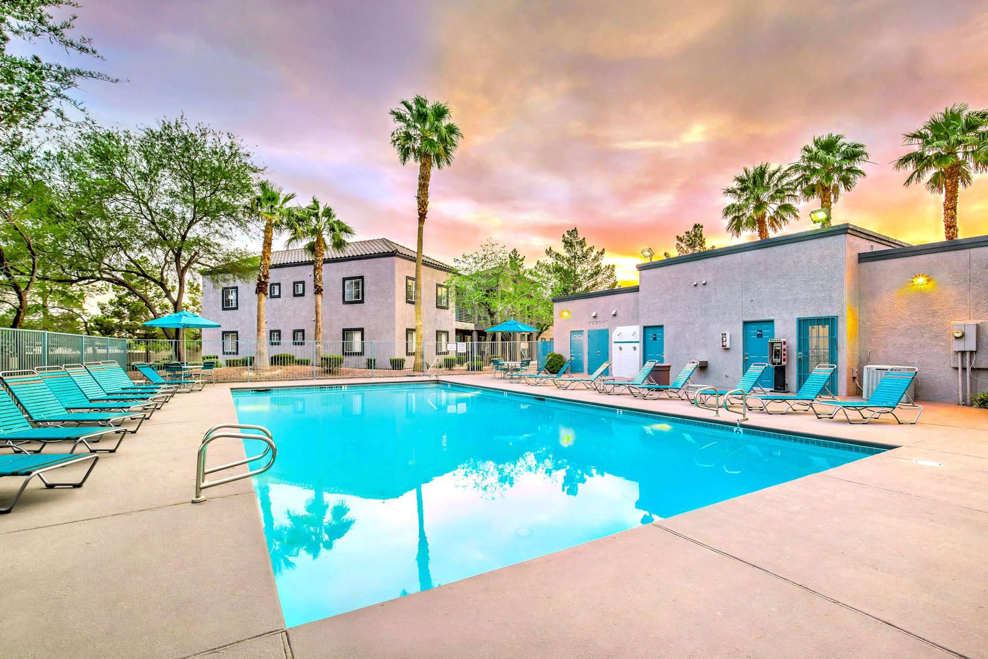 A spacious pool area with lounge chairs and palm trees under a colorful sky at sunset.