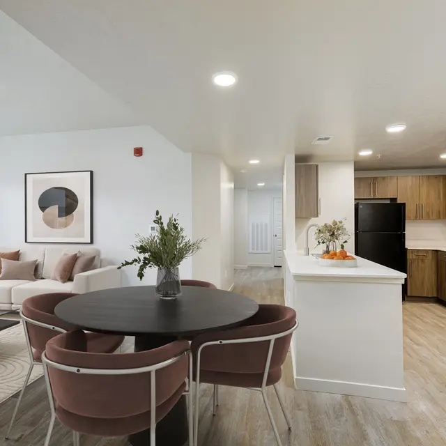 A modern apartment interior featuring a living area with a white sofa and decorative cushions, a circular dining table surrounded by pink chairs, and a sleek kitchen with black appliances and wooden cabinets.