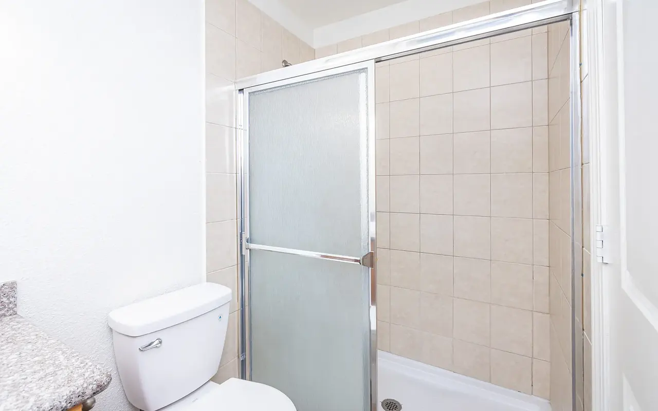 A modern bathroom featuring a shower with a sliding glass door and a white toilet. The walls are tiled in light beige, and there's a small counter with a sink in the room.