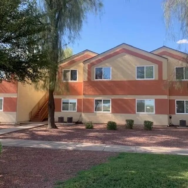 A two-story apartment complex with orange and beige siding, surrounded by green grass and trees. There are three trees and a pathway leading to the entrance, along with decorative bushes in front.