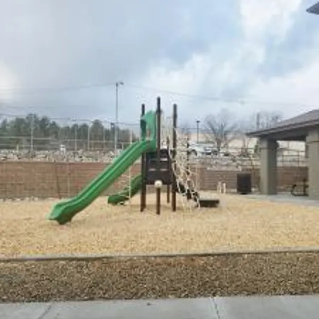 A playground with a green slide and climbing structure surrounded by gravel, seen from a distance with a cloudy sky in the background.
