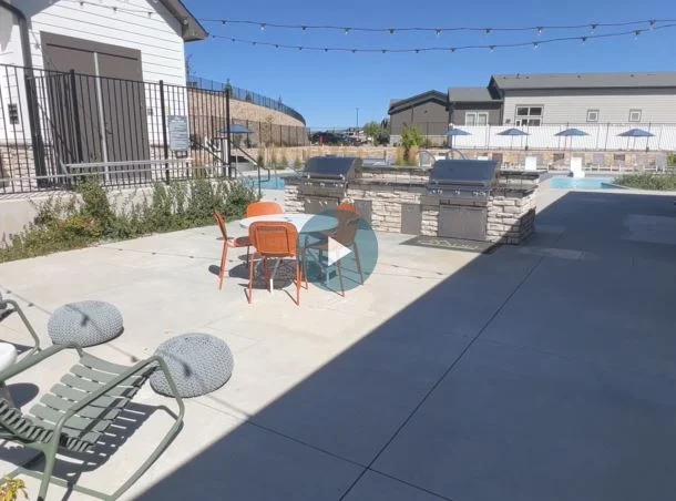 An outdoor gathering area next to a pool, featuring a stone grill, metal chairs, and a nice view of the surrounding greenery and buildings.