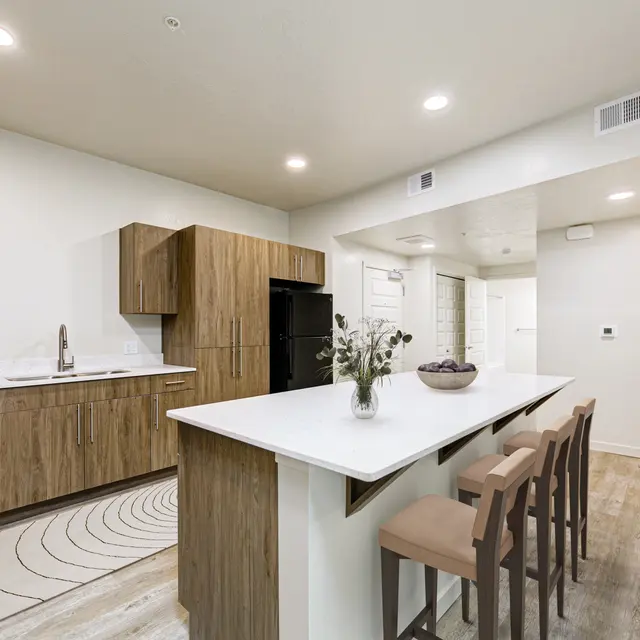 A modern kitchen featuring a large island with seating, wooden cabinetry, and white countertops. There is a black refrigerator and a minimalist design with soft lighting.