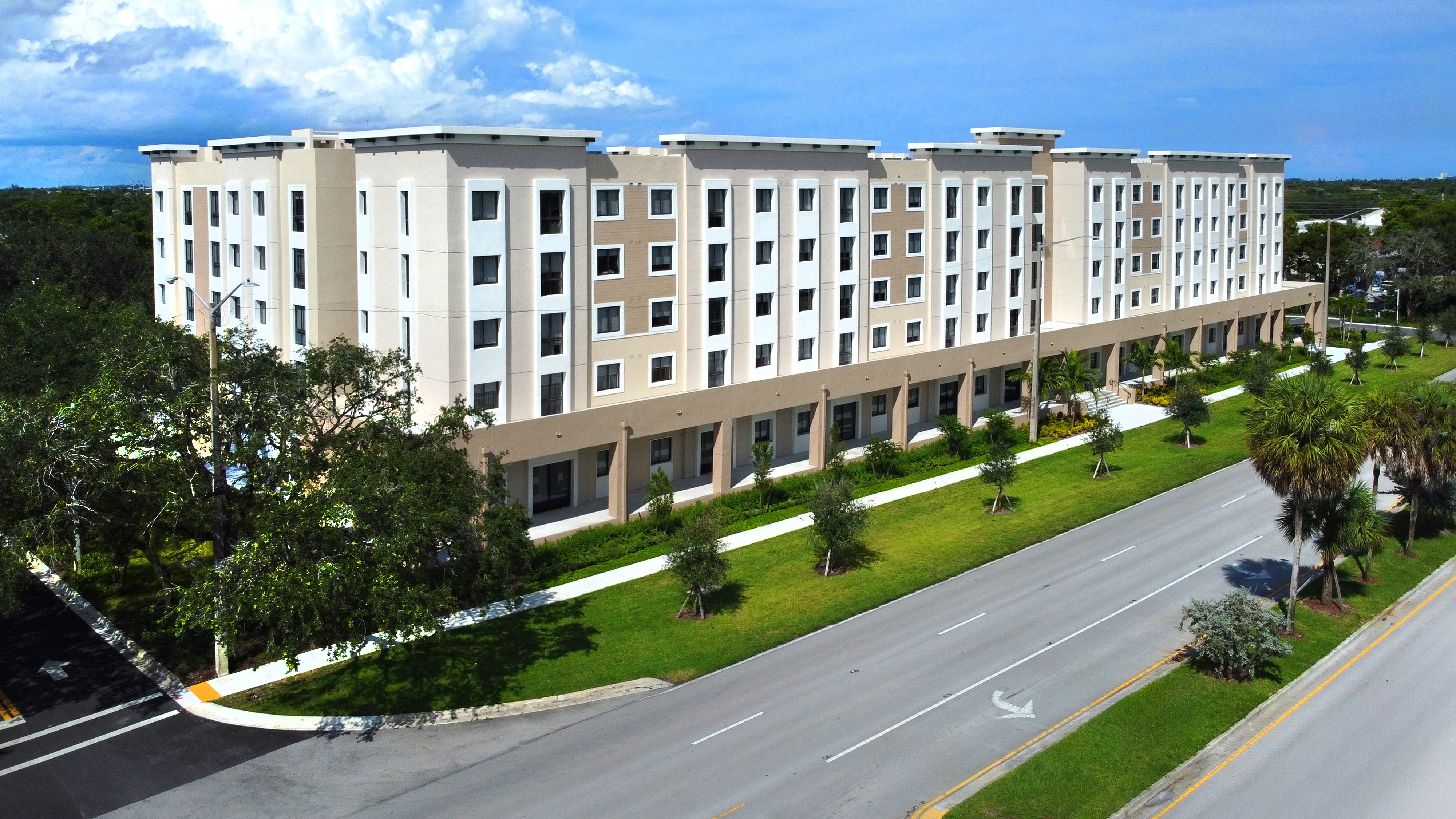 A modern four-story apartment building with a light-colored facade situated along a tree-lined street.