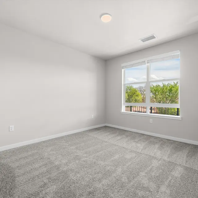 An empty bedroom with light gray carpet, white walls, and a large window allowing natural light.
