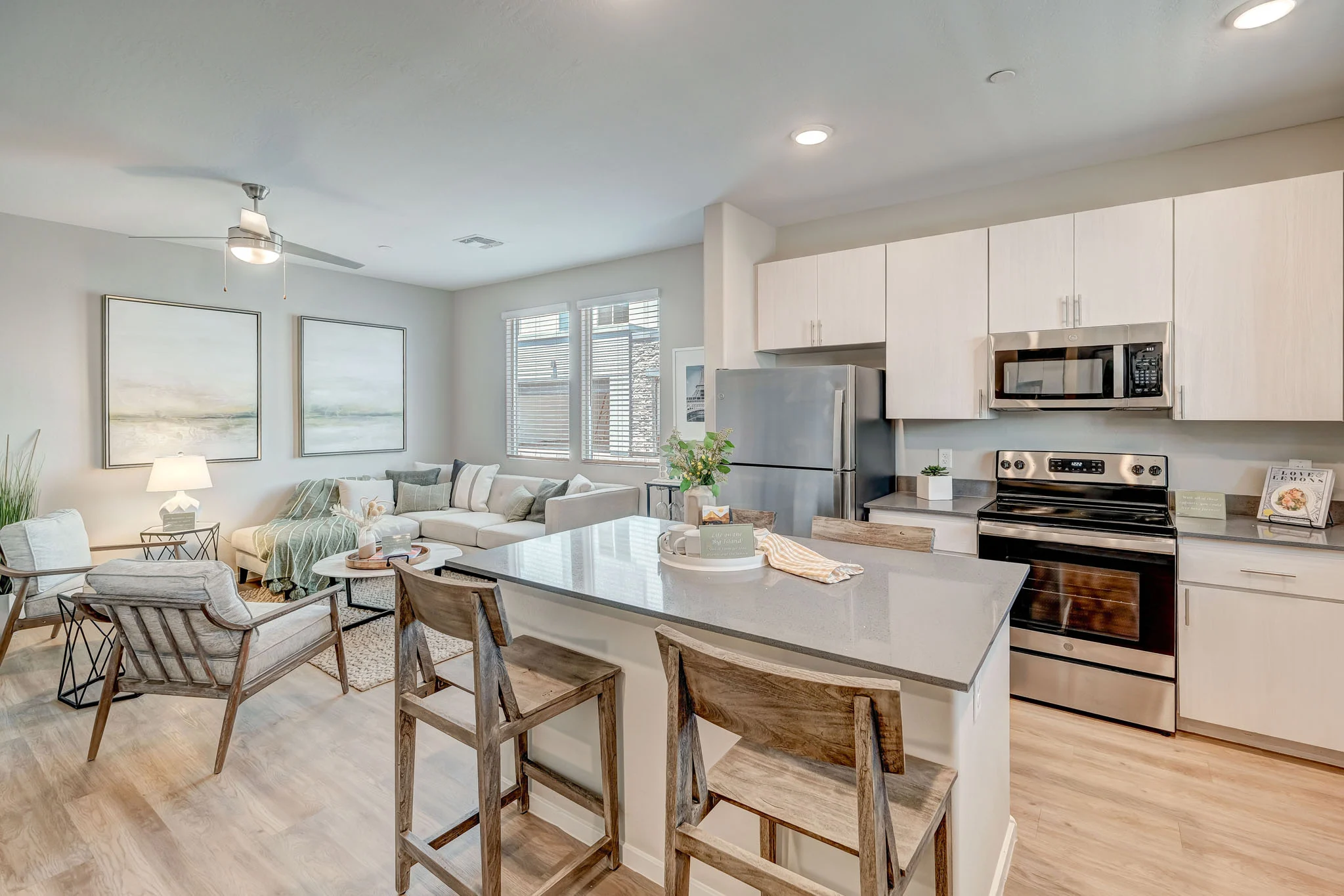 A modern kitchen and living area with a spacious layout, featuring a gray countertop, silver appliances, and a cozy seating area with light-colored furniture.