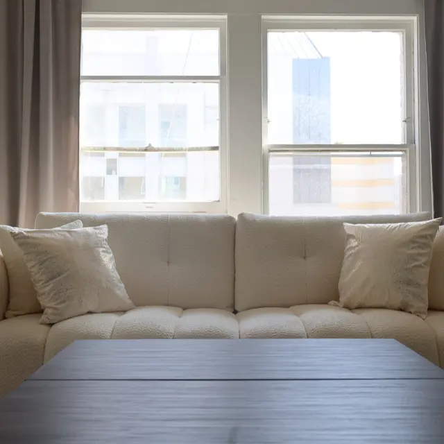 A light-colored sofa with several cushions is positioned in front of two windows with sheer curtains. A wooden coffee table occupies the foreground, enhancing the modern aesthetic of the room. Plants are visible on either side of the sofa, adding a touch of greenery.