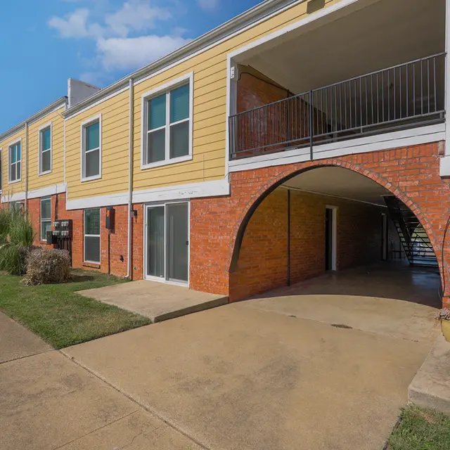 Exterior view of a multi-unit residential building with brick and yellow siding, featuring arches and covered parking.