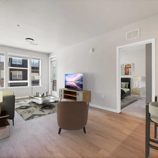Modern Apartment Living Room A modern living room featuring a gray sofa, two chairs, a coffee table, and a wall-mounted TV. Large windows allow natural light, with a view of an outdoor area. The space is airy and decorated with neutral colors and a patterned rug. To the right, there's an open doorway leading to another room.