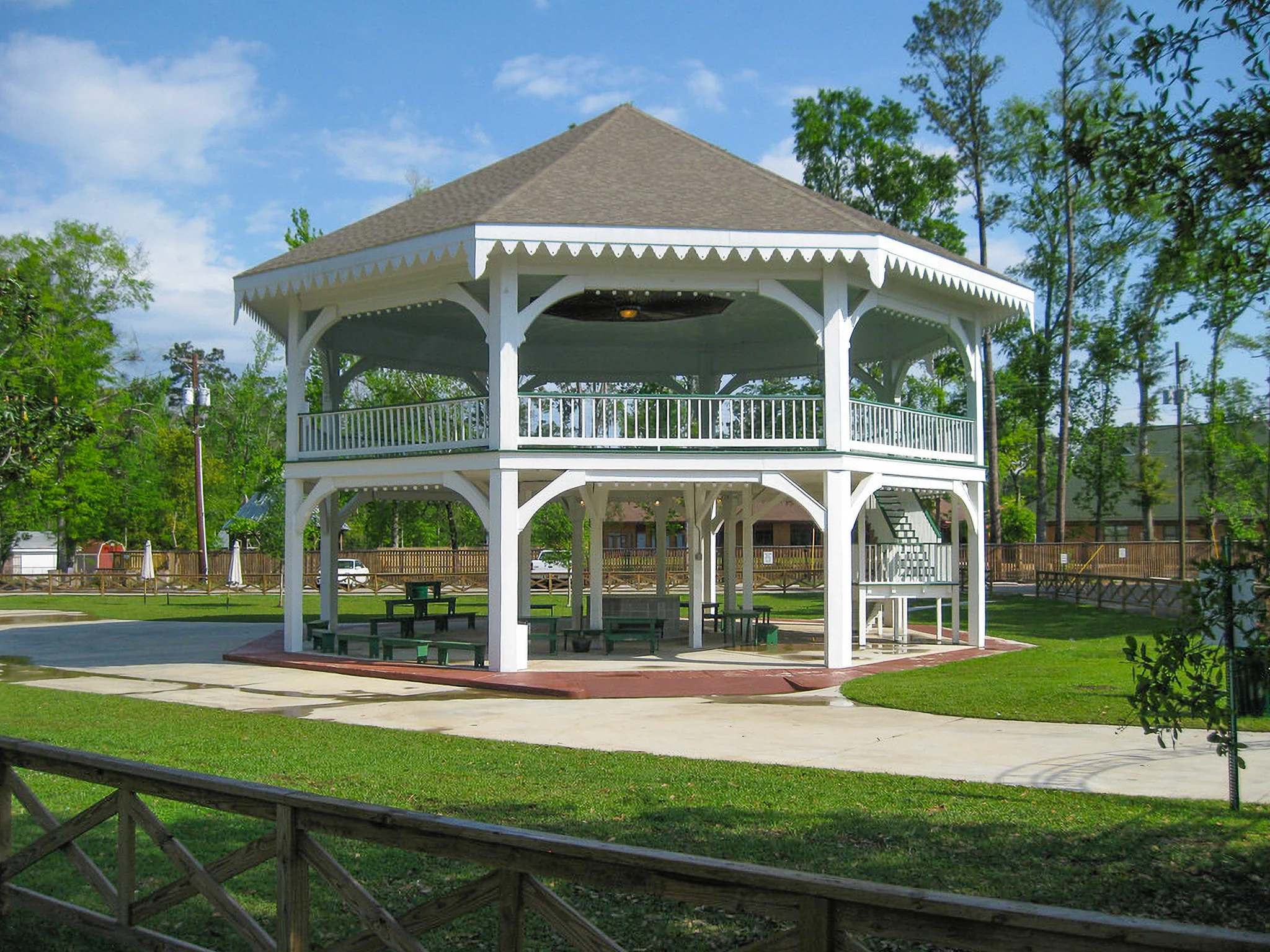 A white wooden gazebo with a thatched roof situated in a park, surrounded by greenery and paved pathways.