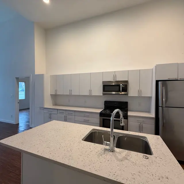 A modern kitchen featuring white cabinetry, stainless steel appliances, and a central island with a sink. The space is bright and spacious with high ceilings and wooden flooring.