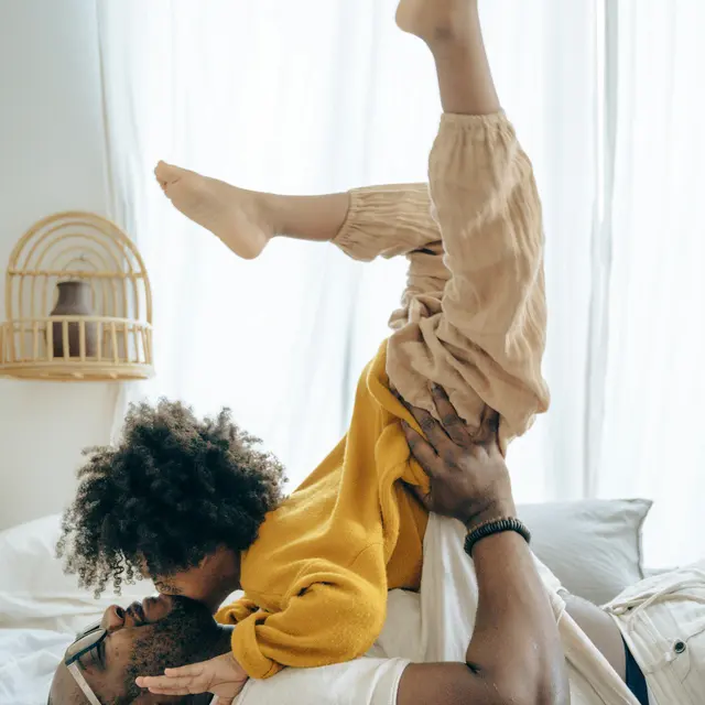 Parent and Child Playtime A parent playfully lifting a child upside down while lying on a bed. The child has curly hair and is wearing a yellow sweater, while the parent is shirtless and wearing a bracelet. Natural light fills the room.