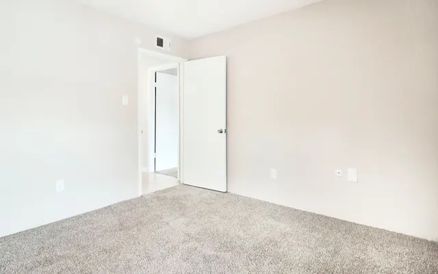 A minimalist empty room with beige walls and a light gray carpet. A door is slightly ajar on the left side, revealing another doorway. There are no furnishings or decorations in the space.