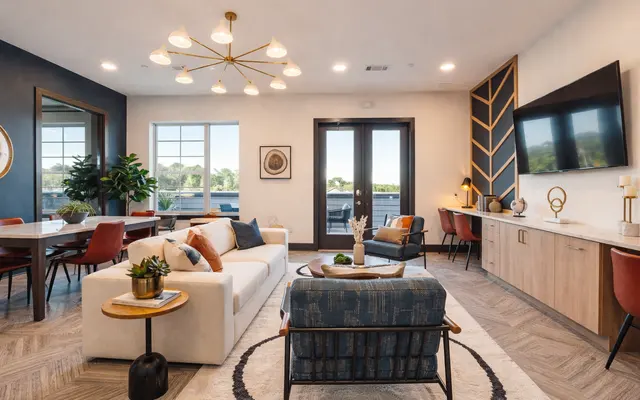 Contemporary Living Room Design A modern living room featuring a neutral color scheme with a stylish couch, decorative pillows, a round side table, and a patterned rug. The room has large windows providing natural light, a dining space to the left, and a wall-mounted television. Decorative plants and minimalist furniture enhance the contemporary aesthetic.