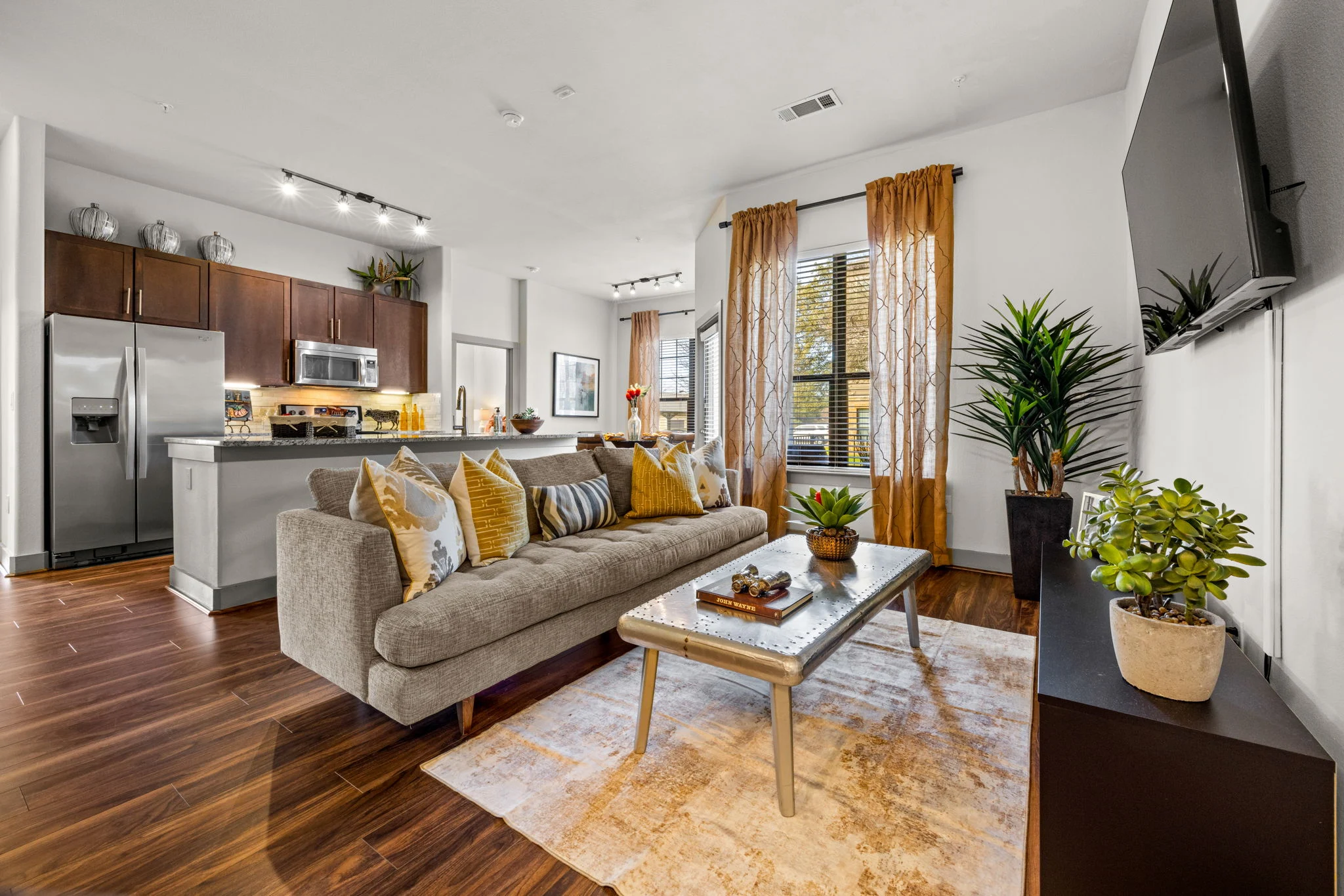 A stylish modern apartment living room featuring a gray sectional sofa adorned with patterned pillows. The space includes wooden flooring, a glass coffee table, and decorative plants. In the background, there's a kitchen area with dark wooden cabinets and stainless steel appliances, complemented by large windows with light curtains.