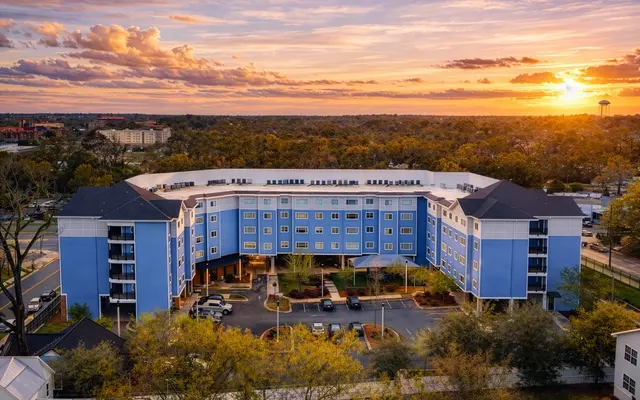 Aerial view of a blue hotel building with sunset sky, surrounded by trees and parking area.