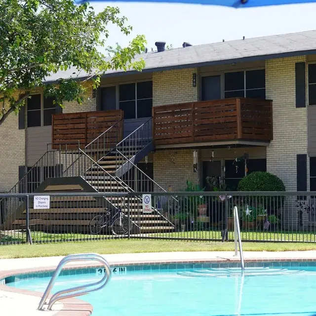 A view of a swimming pool in front of an apartment complex, with a staircase leading to the second floor and wooden balconies.