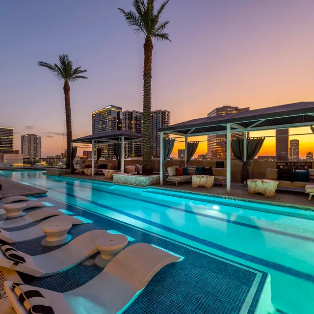 A luxury rooftop pool at sunset with lounge chairs and cabanas, palm trees, and a city skyline in the background.