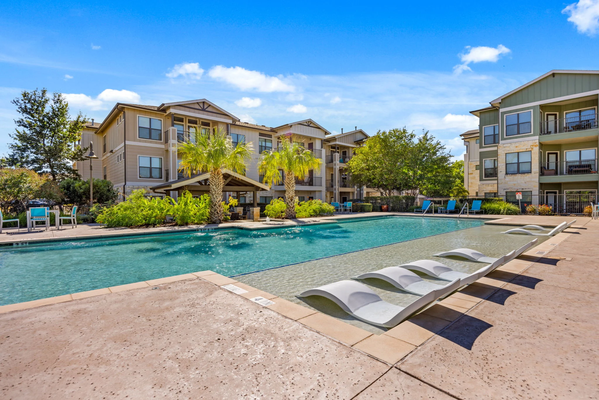 A spacious apartment complex pool area featuring a large swimming pool, lounging chairs, palm trees, and surrounding greenery under a blue sky.